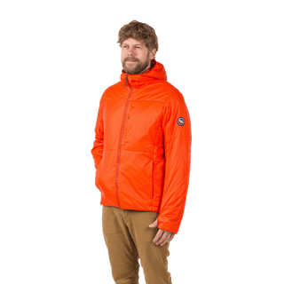 A bearded man with curly hair wears the M's Barrows Jacket by Big Agnes in orange, made from recycled Pinneco, with a circular sleeve patch and tan pants, standing against a plain white background.