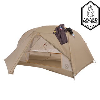 A light brown, dome-shaped Big Agnes Tiger Wall UL2 Bikepack Solution Dye tent stands with its front flap open, revealing the interior. A dark jacket with colorful patches hangs on top, and an Award Winning badge appears in the upper right corner.